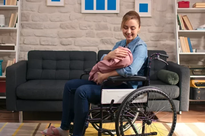 A woman who uses a wheelchair holding a baby in her arms in a living room.